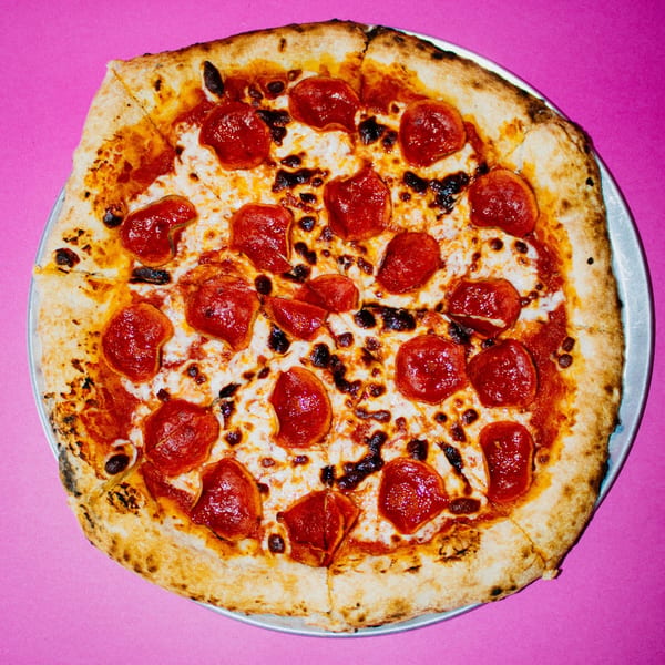 A whole pepperoni pizza from Tank's Pizza, with a golden crust and melted cheese, on a pink background.